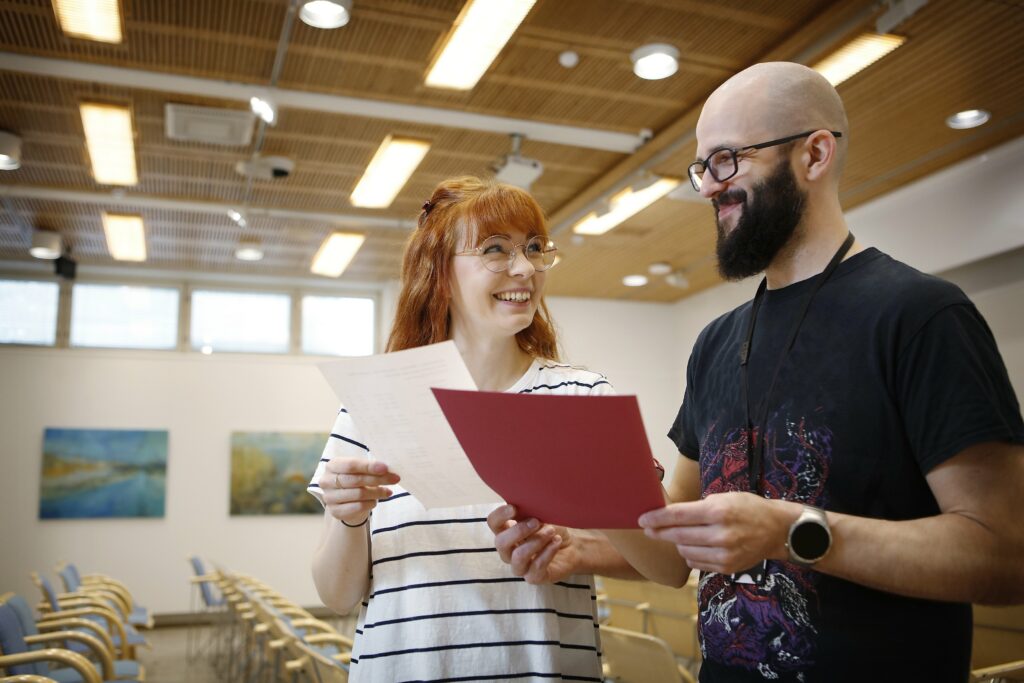 Nainen ja mies seisovat vierekkäin, pitelevät käsissään papereita ja katsovat hymyillen toisiaan.